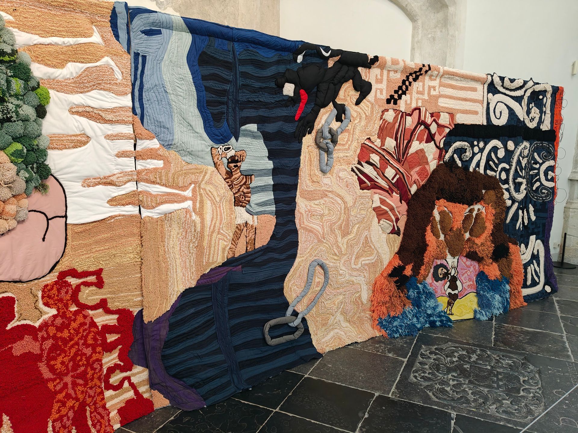 Een kleurrijk mozaïekmuurschilderij met abstracte figuren en gezichten op een achtergrond van tegelvloer.