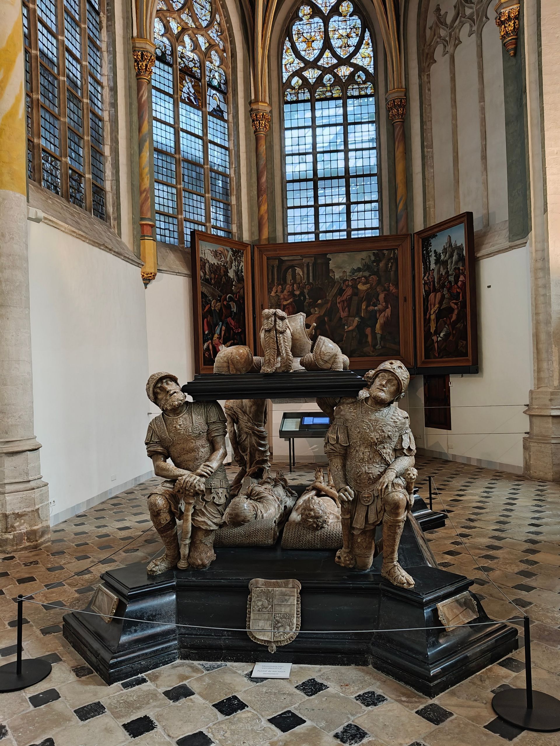 Barokke beeldhouwkunst in een kapel, met sierlijke figuren rond een zwart voetstuk onder hoge glas-in-loodramen.