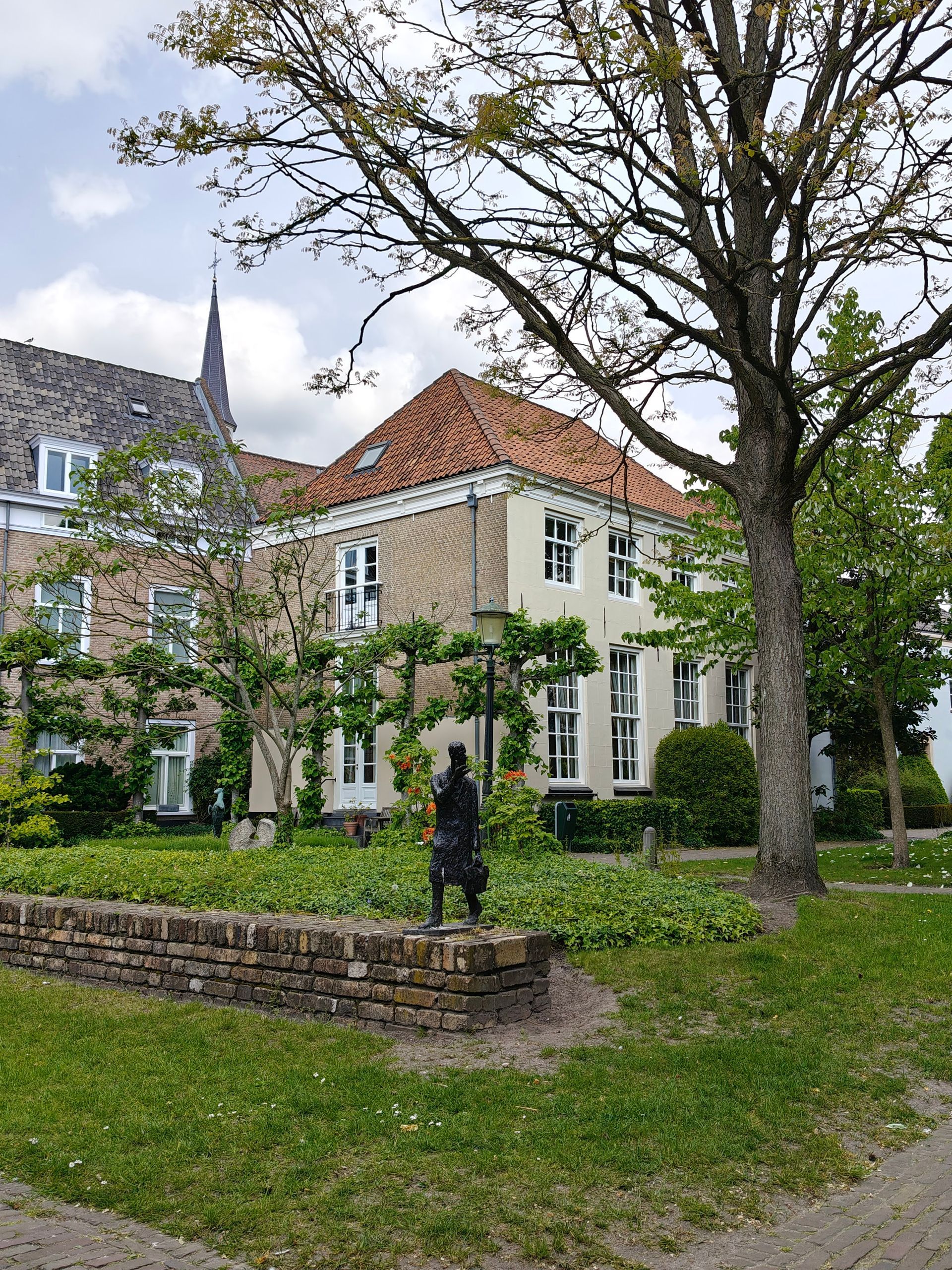 Stenen huis met rood pannendak en grote bomen in een groene tuin, naast een lage bakstenen muur.
