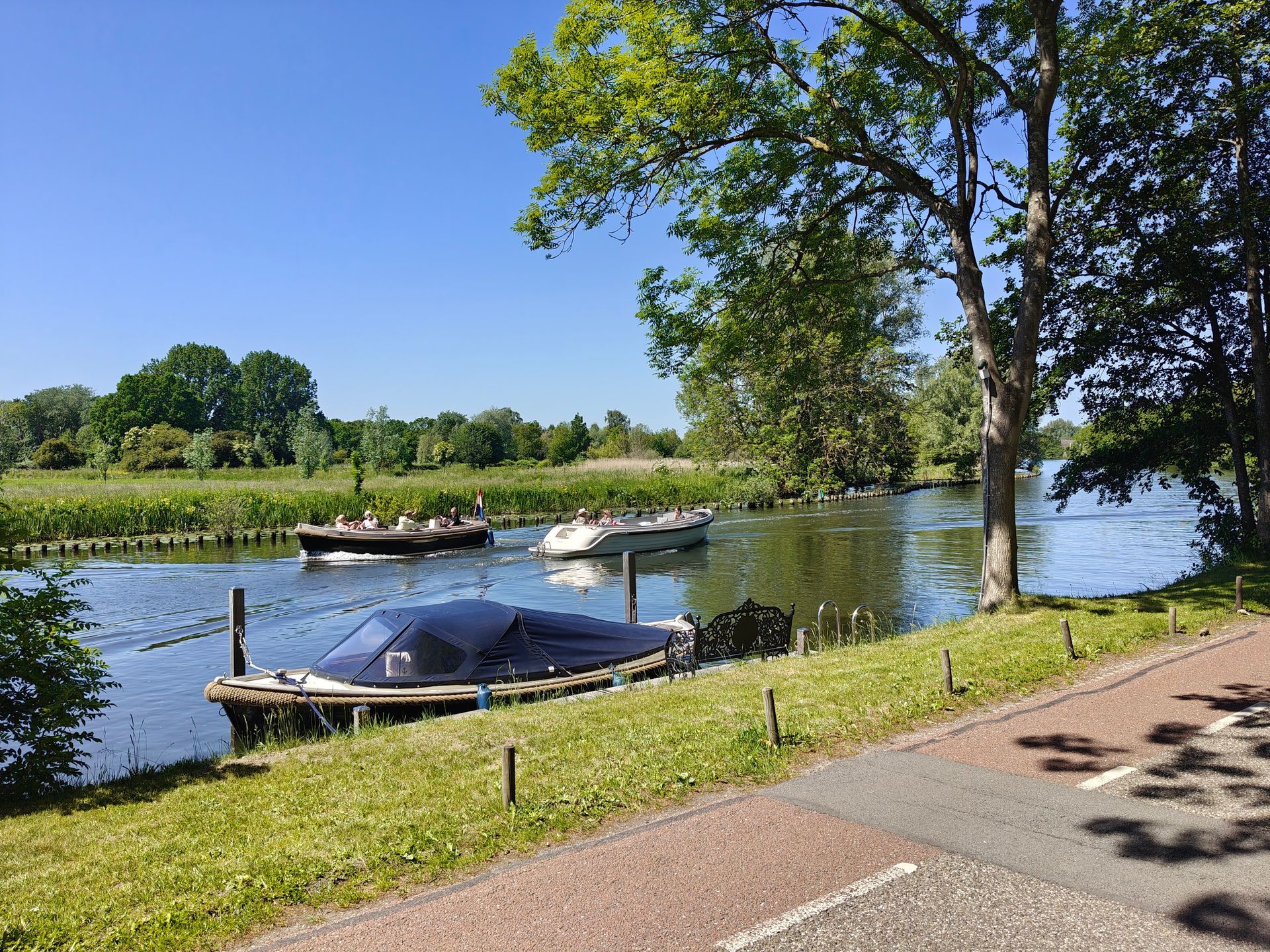 Kleine bootjes liggen aangemeerd aan een rustige rivier naast een graspad en bomen op een zonnige dag.