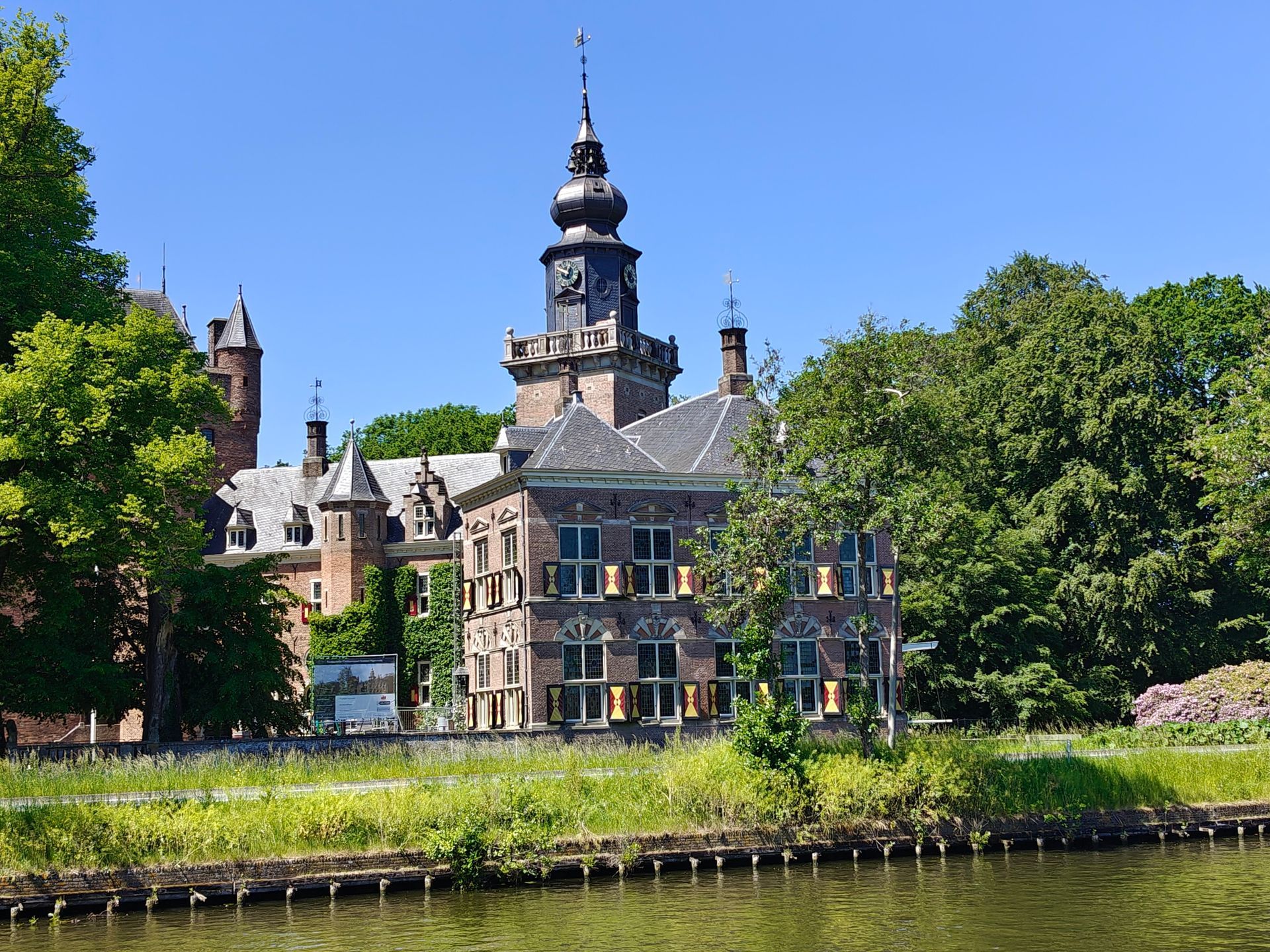 Historisch stenen gebouw met een toren naast een kanaal, omgeven door groene bomen onder een blauwe hemel.