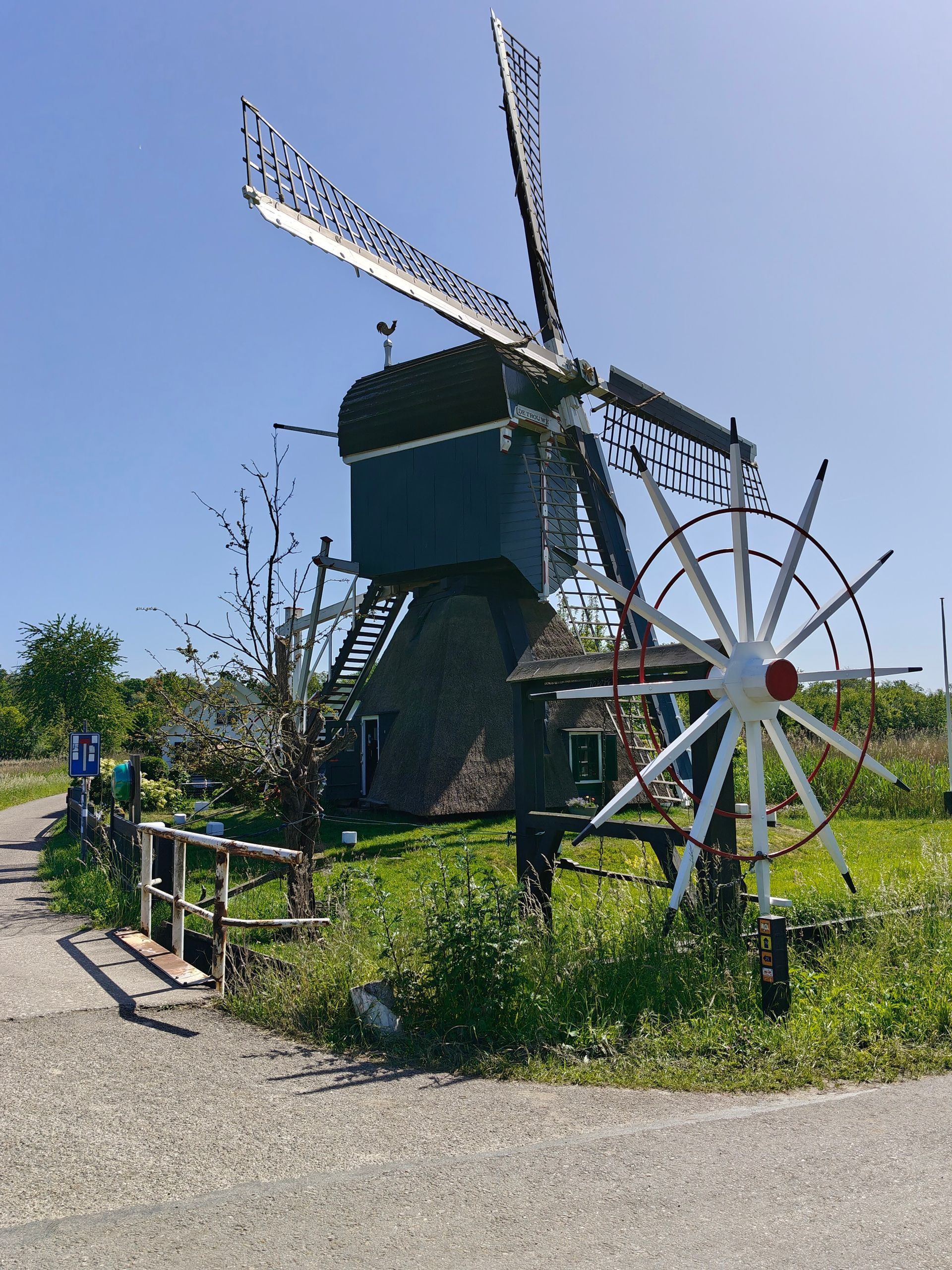 Historische windmolen met witte wieken en een groot wiel naast een grindpad in een zonnig veld.