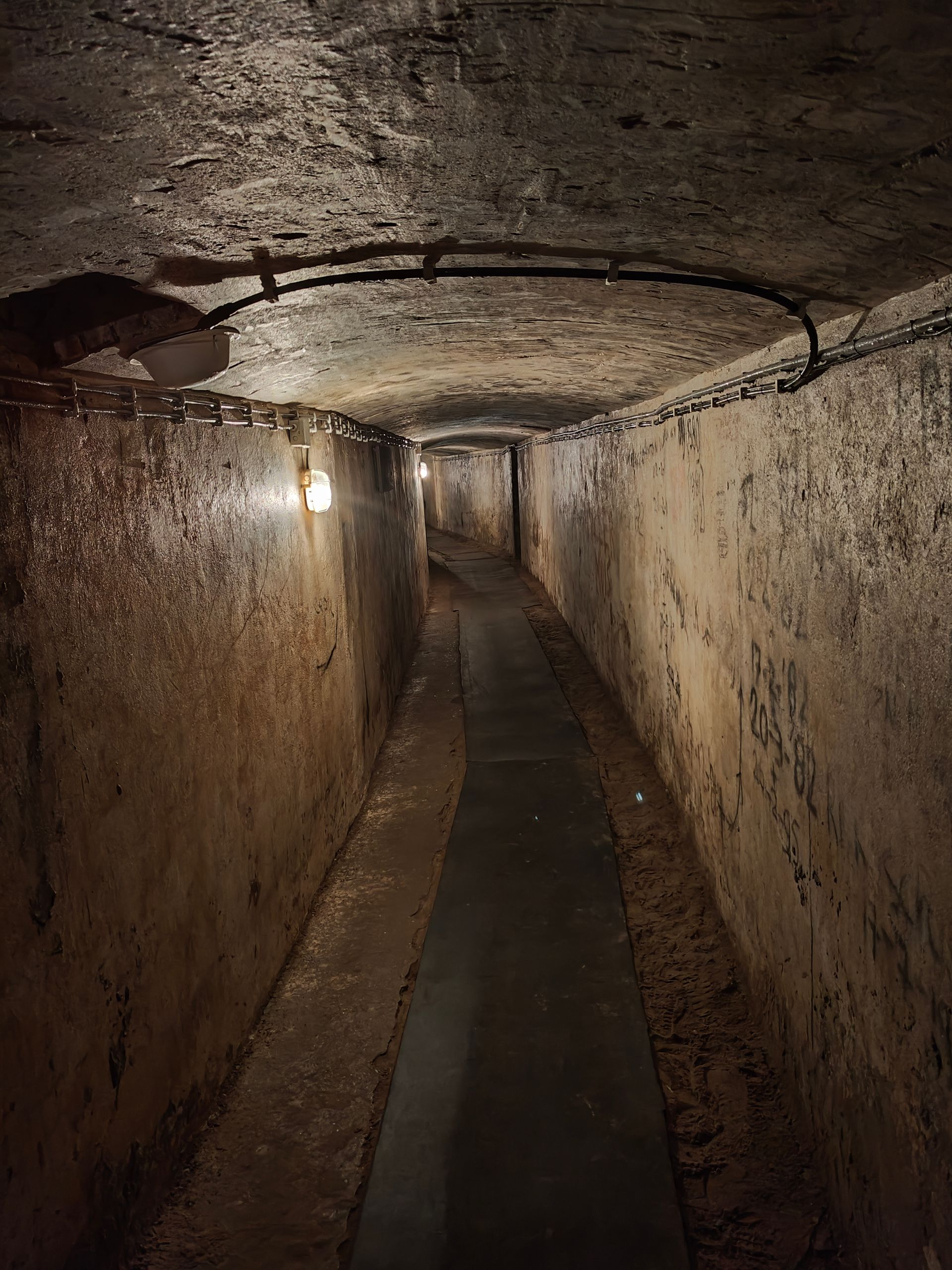 Smalle ondergrondse stenen tunnel met een betonnen loopbrug en een enkele lamp die de doorgang verlicht.