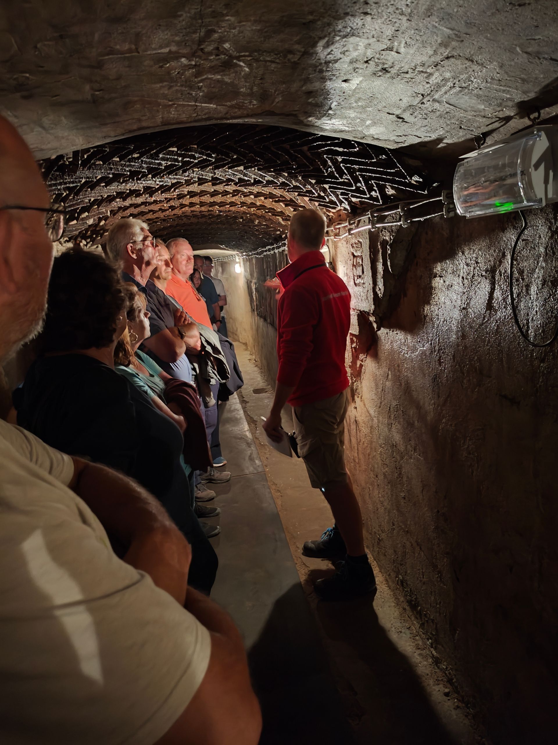 Een rondleidingsgroep loopt door een smalle, schemerige ondergrondse tunnel met rotswanden en plafondverlichting.