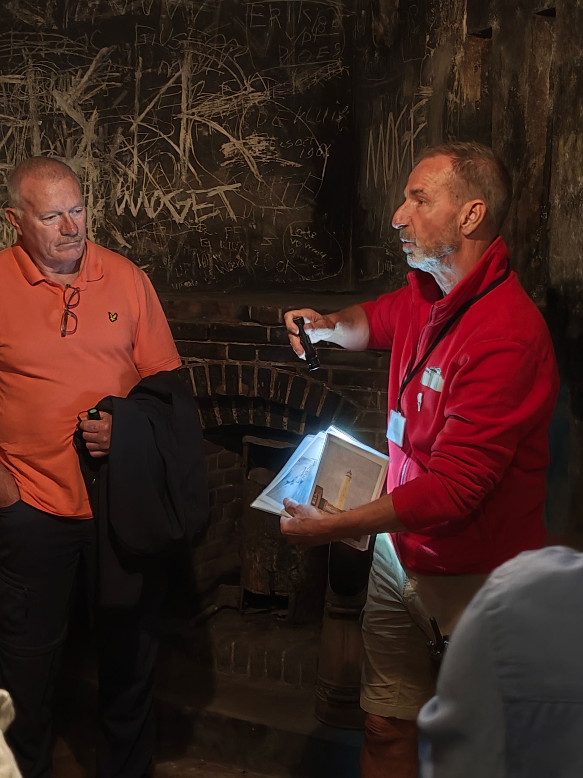 Twee mensen in een schemerige, rustieke kamer, de een in rood gebarend met een verlicht tablet, de ander in oranje luisterend