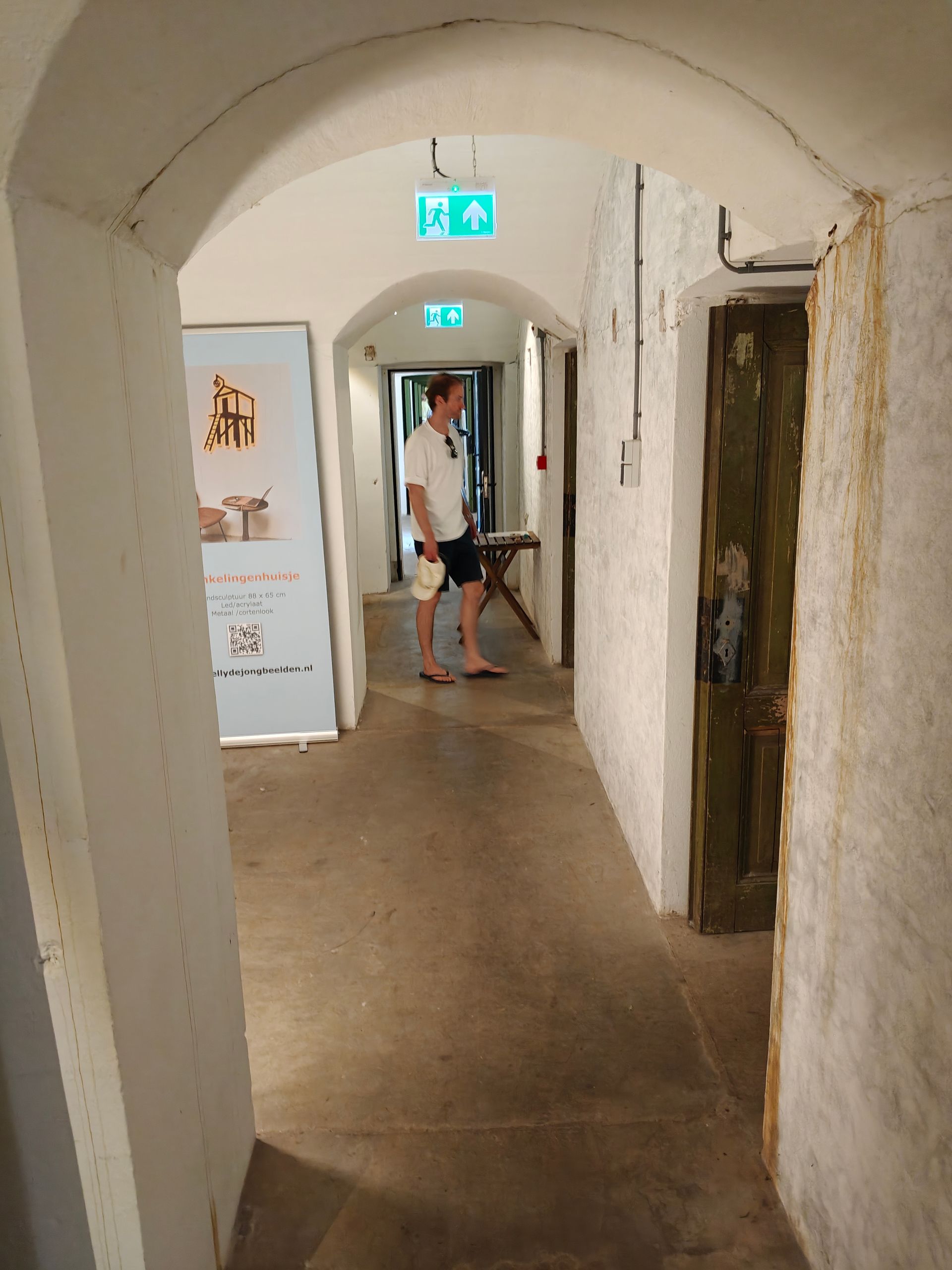 Smalle, gewelfde gang met witte muren, een persoon die bij een deuropening staat en groene nooduitgangborden boven het hoofd.