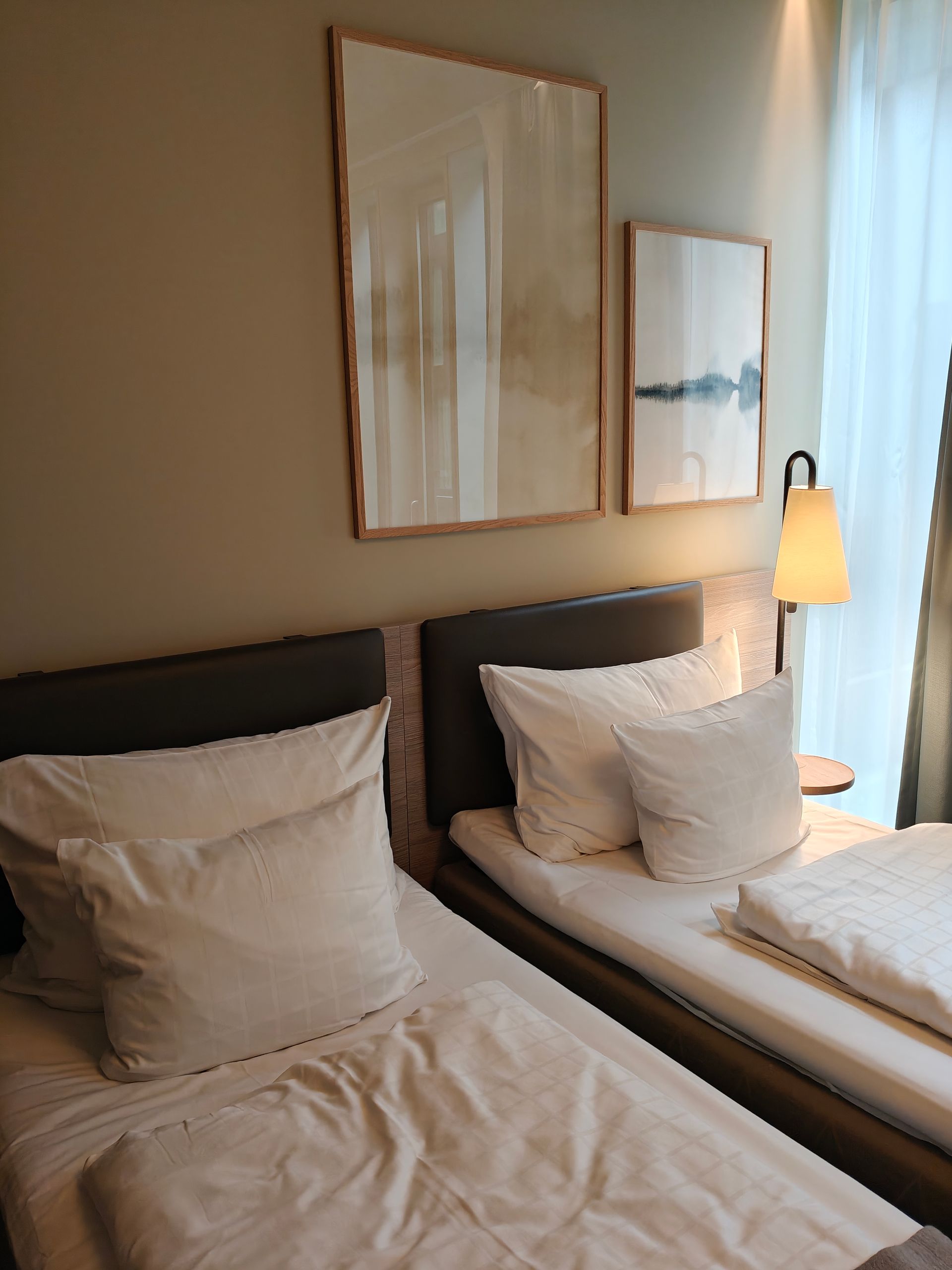 Twee keurig opgemaakte eenpersoonsbedden in een lichte, moderne hotelkamer met een nachtlampje en wanddecoratie.