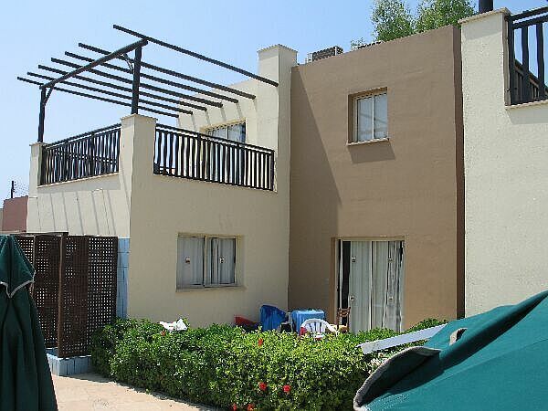 Een beige huis met twee verdiepingen, een balkon, een pergola en groene struiken ervoor, onder een heldere hemel.