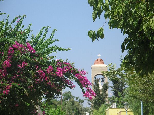 Bloeiende roze bougainvillea en groene bomen omlijsten een witte toren met een gouden koepel onder een blauwe hemel.