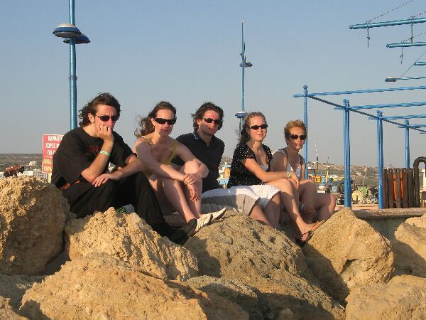Vijf mensen zitten op rotsen op een strand met blauwe palen en een heldere hemel op de achtergrond.