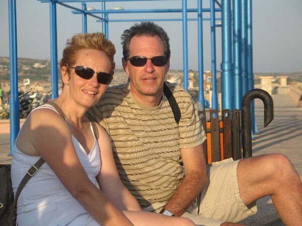 Twee mensen met zonnebrillen zitten samen buiten bij een blauwe reling, met een zonnig strand op de achtergrond.