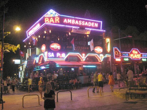 Een bar en club met neonverlichting 's nachts, met een menigte buiten en opvallende borden.