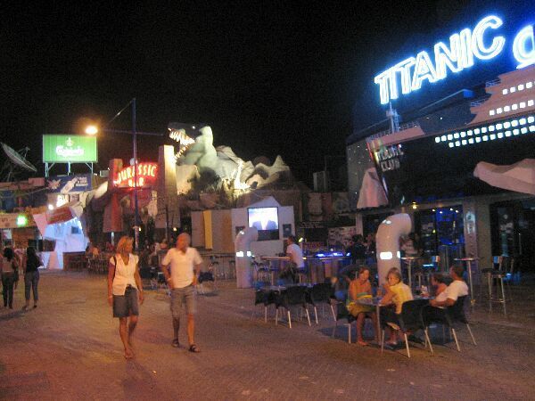 Een terras en promenade 's avonds met neonreclames, wandelende mensen en gasten aan tafels.
