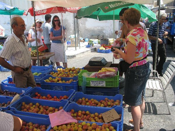 Een marktkraam in de openlucht met kleurrijke producten in blauwe kratten en twee mensen die onder parasols rondkijken.