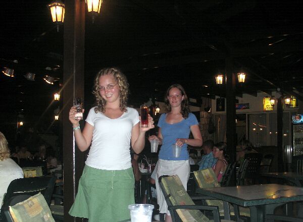 Twee vrouwen met drankjes in hun handen op een schemerig verlicht terras 's avonds.