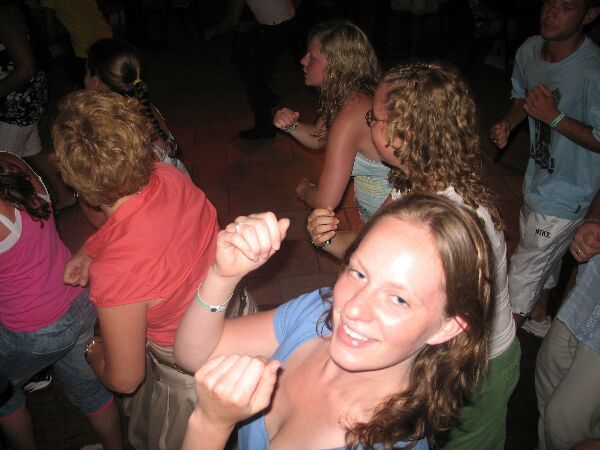 Mensen dansen in een schemerig verlichte menigte, met een lachende vrouw in een blauw shirt op de voorgrond.