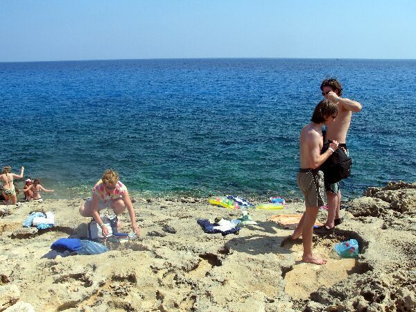 Mensen ontspannen op een rotsachtig strand aan de blauwe oceaan, met zwemmers en handdoeken langs de kust.