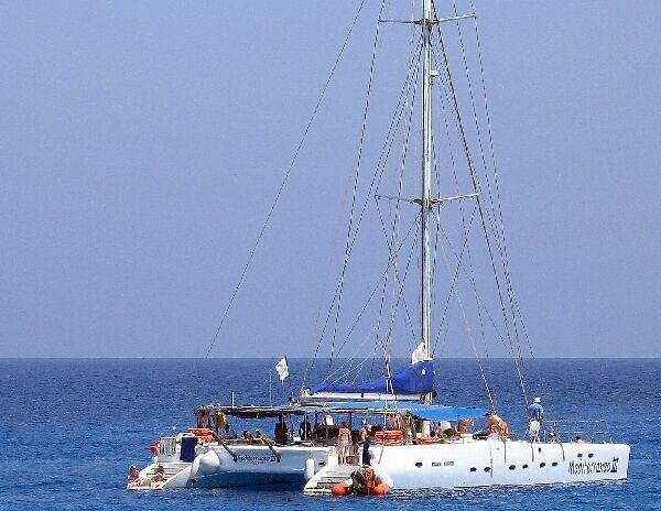 Zeilboot/catamaran met mensen aan boord op een kalme blauwe oceaan onder een heldere hemel.