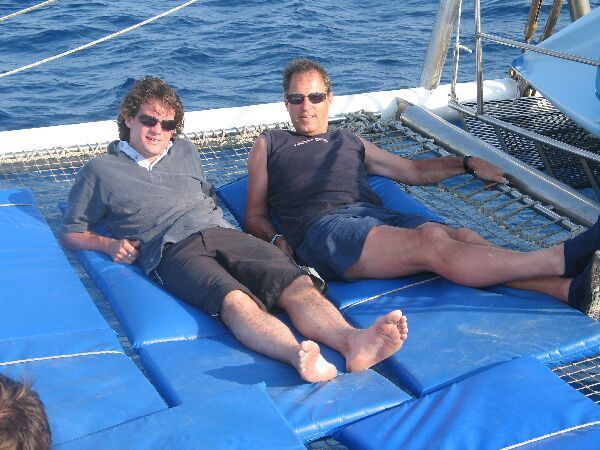 Twee mensen ontspannen op het dek van een blauwe boot met de oceaan op de achtergrond.