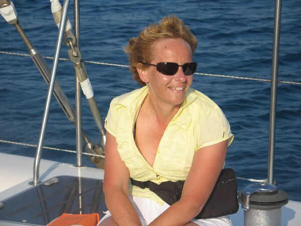 Een vrouw lacht op een zeilboot, met een zonnebril en een geel shirt aan, terwijl ze op het water vaart.
