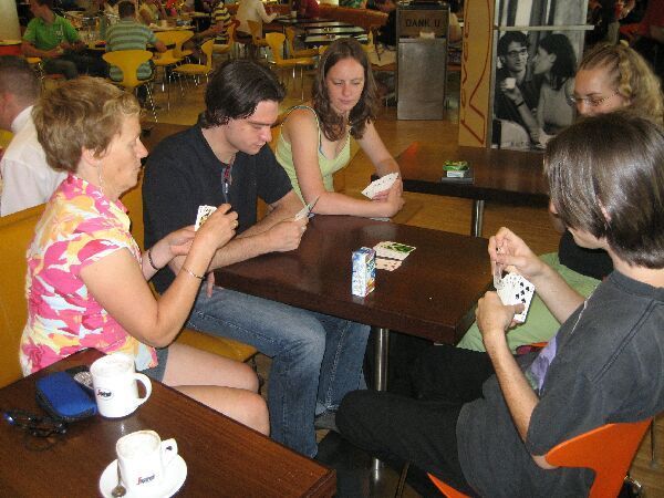Vier mensen spelen kaart aan een cafétafel, met koffiekopjes en een kleurrijk interieur.