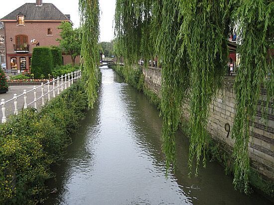 Een smal kanaal, omzoomd met treurwilgen en stenen muren, naast huizen en een wandelpad.