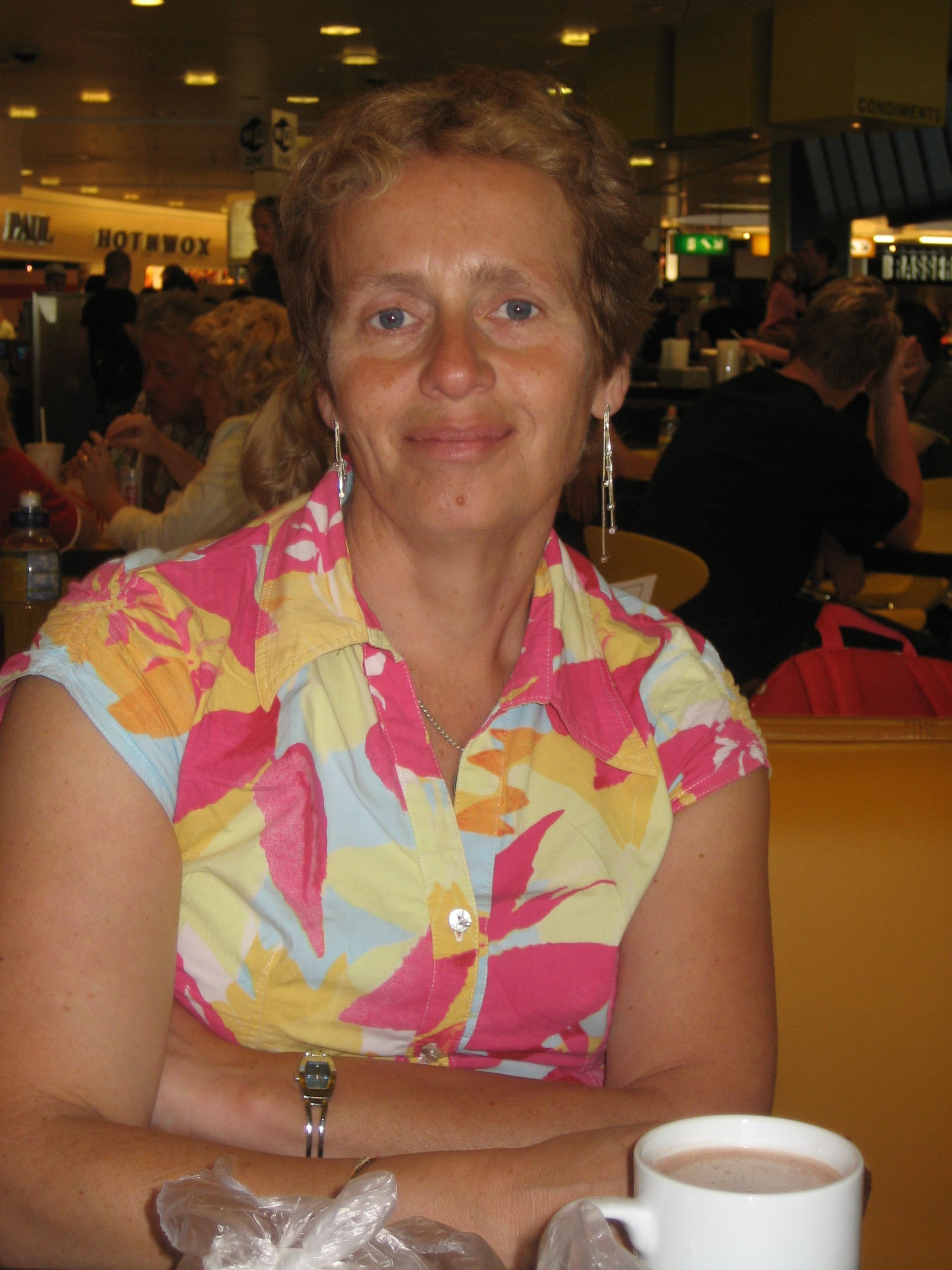 Vrouw in een roze blouse met bloemenprint zit met een kopje koffie aan een tafeltje in een druk café.