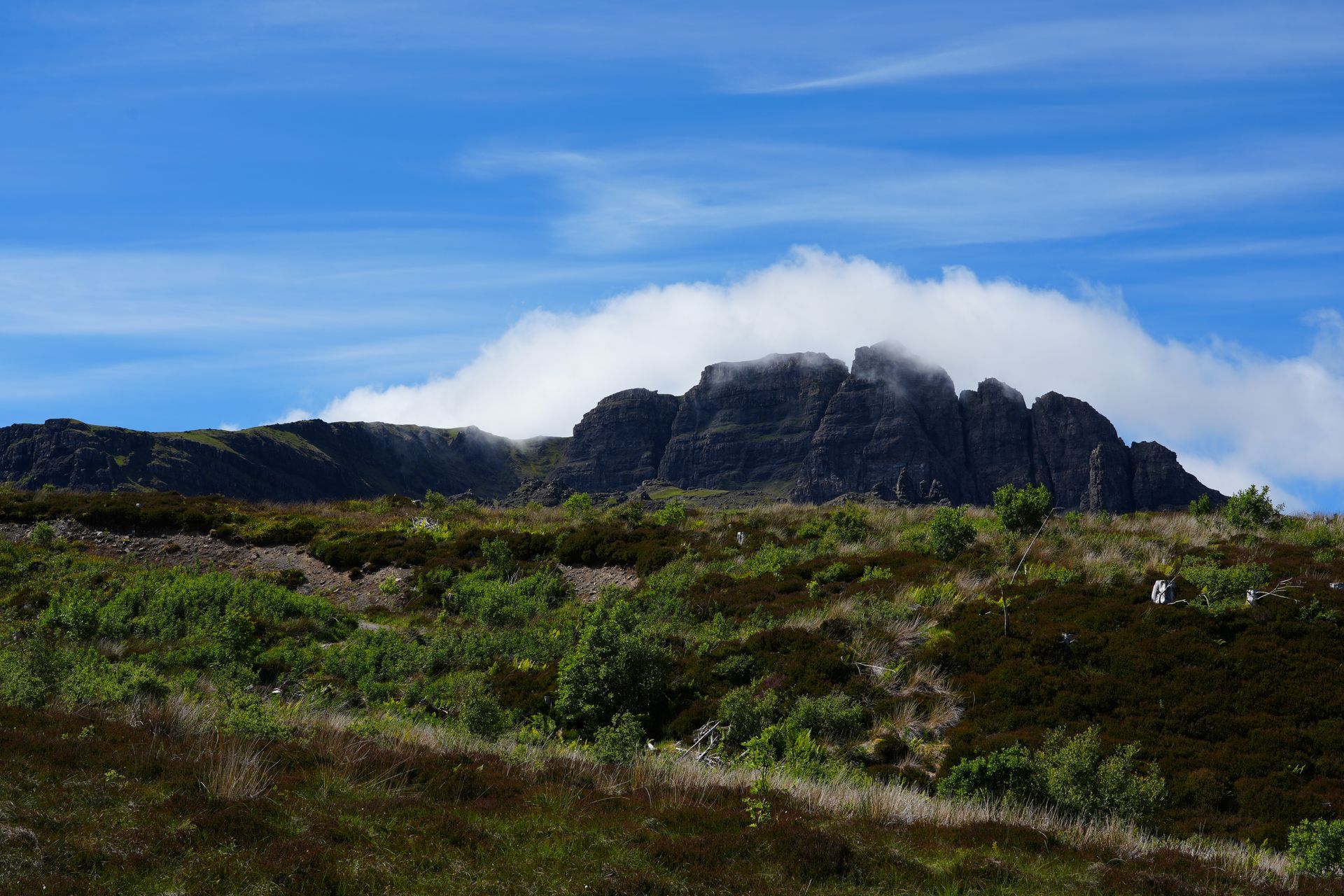 Een rotsachtige bergtop met wolken, groene vegetatie en een blauwe lucht.