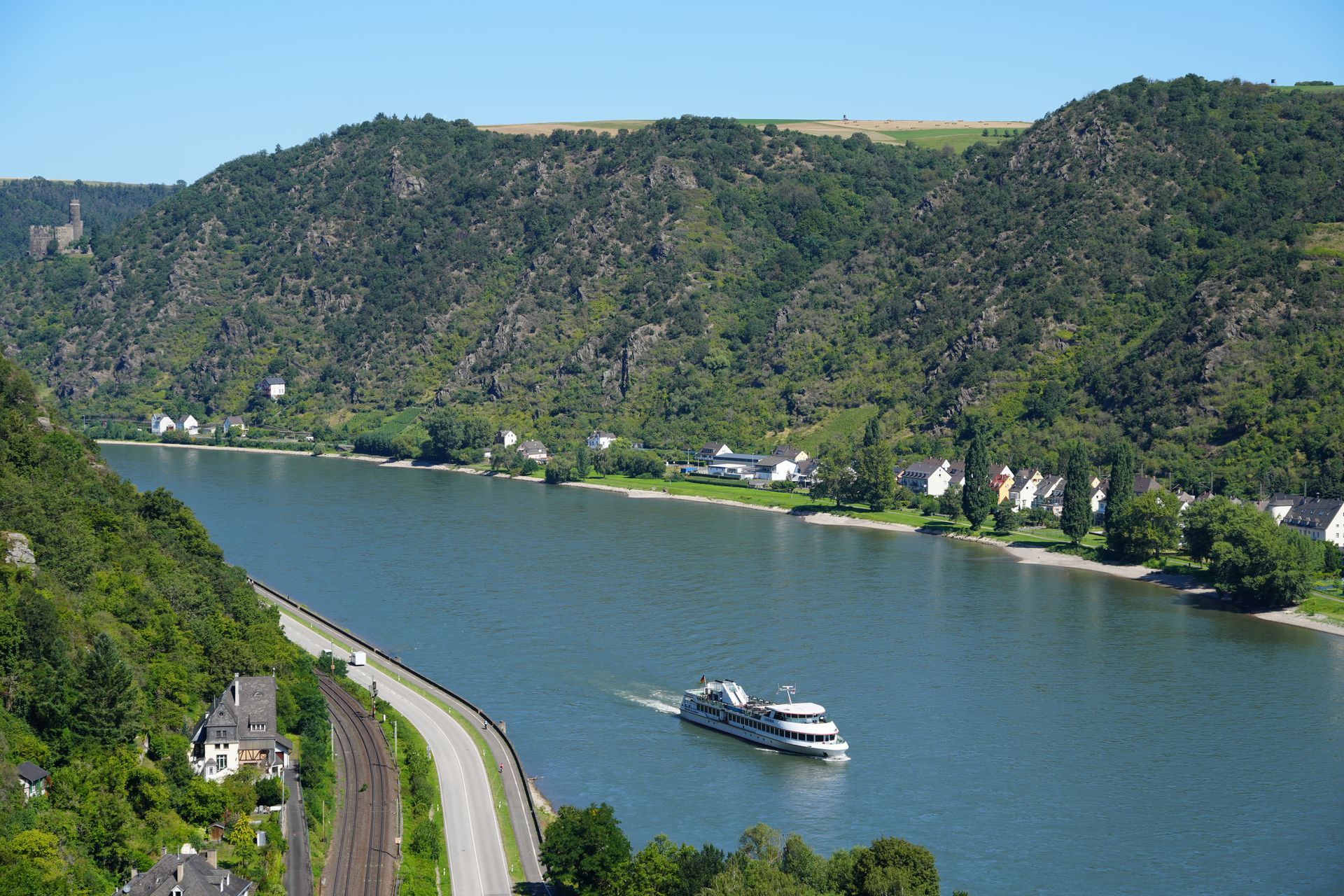 Een riviercruiseschip vaart over een brede rivier, geflankeerd door beboste heuvels en een weg met gebouwen.
