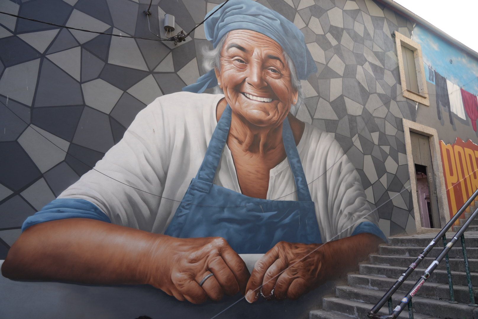 Muurschildering van een lachende vrouw met een blauwe hoofddoek en schort, rustend op een richel. Porto