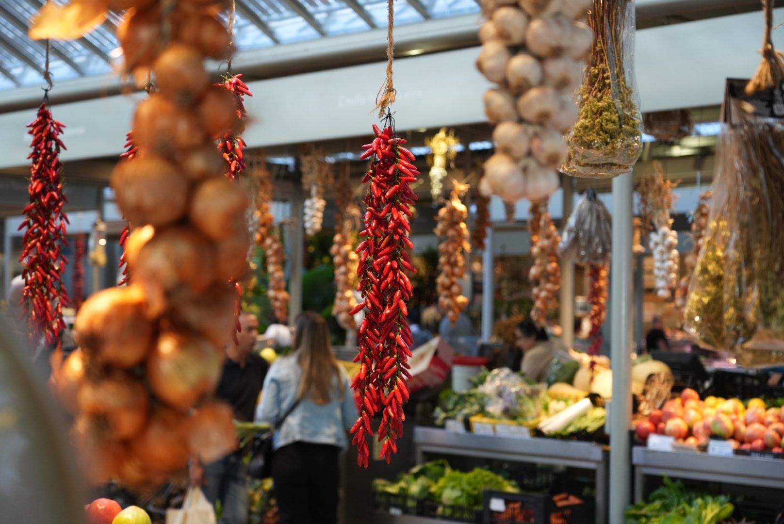 Slingers met uien, rode paprika's, knoflook en kruiden hangen in een marktkraam tussen de winkelende mensen.