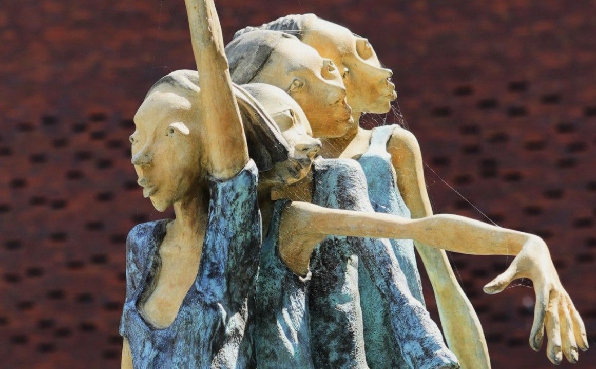 Abstracte sculptuur van drie overlappende figuren in blauw en crème, met één opgeheven arm tegen een rode achtergrond.