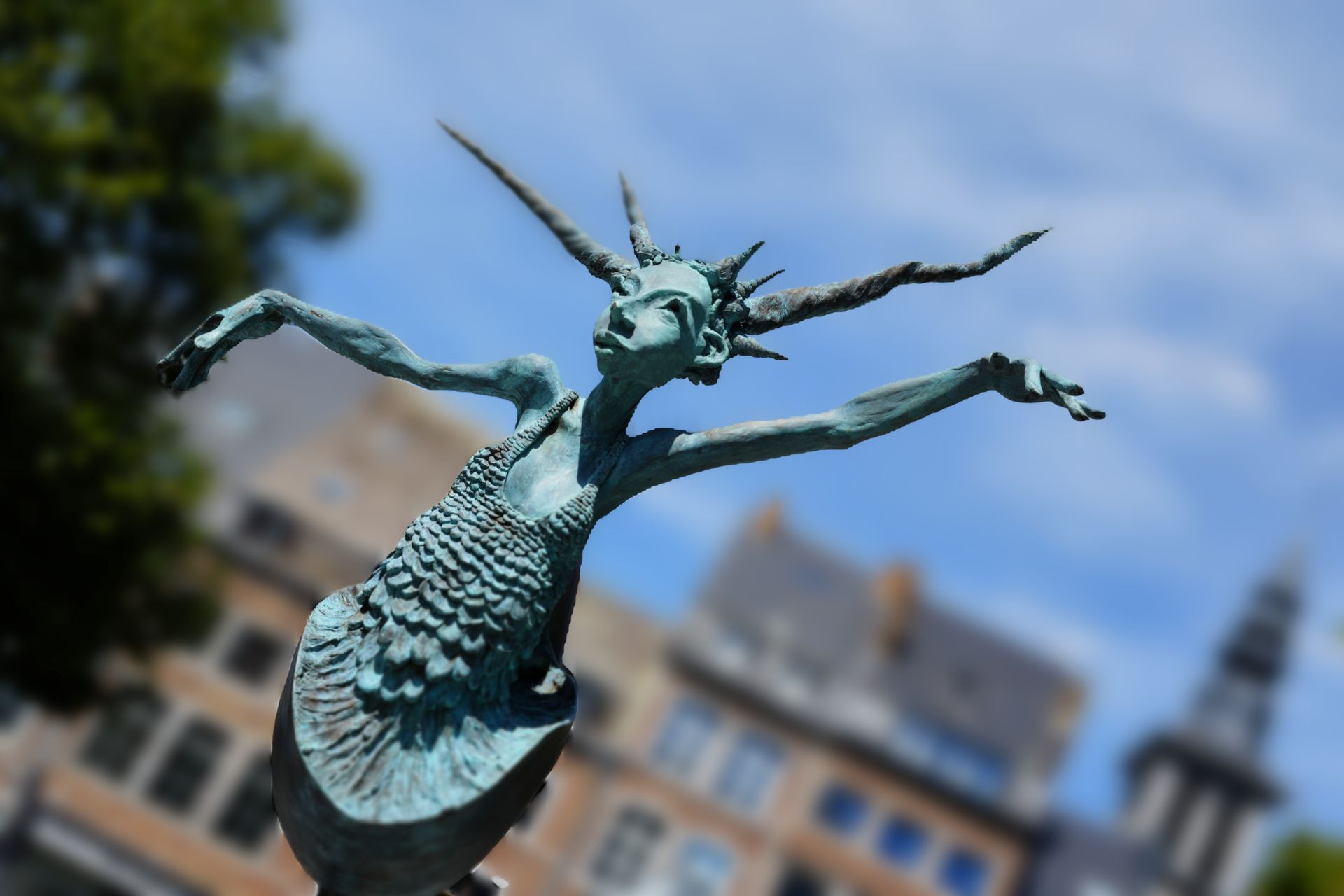 Bronzen beeld met uitgestrekte armen op een stadsplein, met wazige gebouwen en een blauwe lucht op de achtergrond.