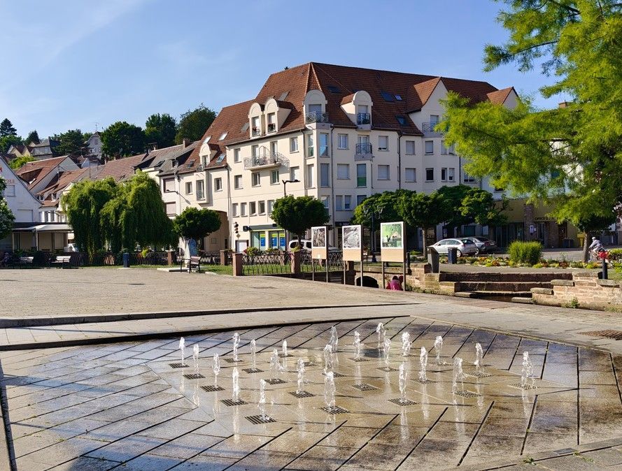 Fontein met waterstralen op een plein; op de achtergrond staan ​​flatgebouwen en bomen onder een blauwe hemel.