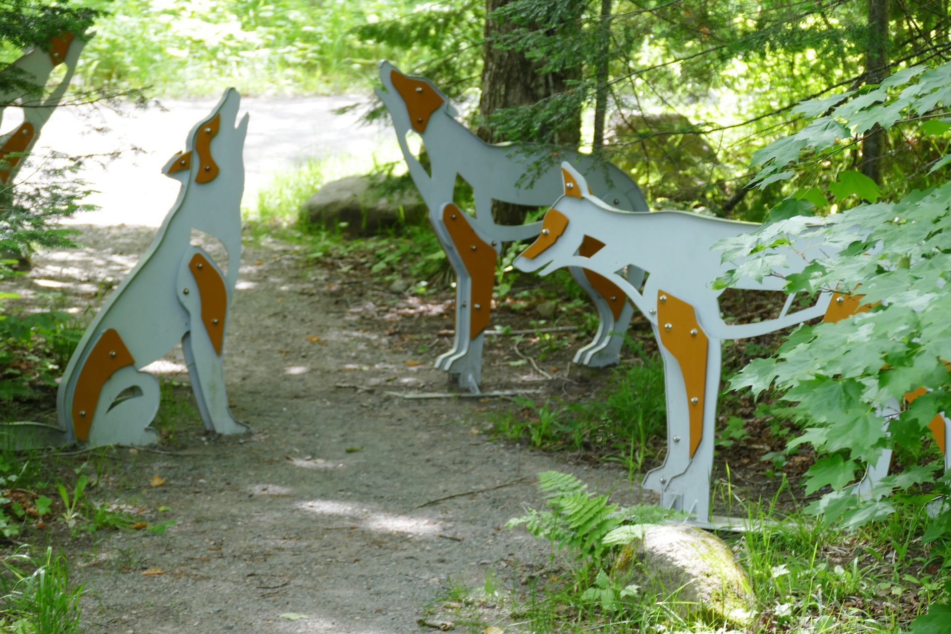 Drie metalen wolfsculpturen in een parkomgeving, waarvan er één huilt, met oranje en grijze details.