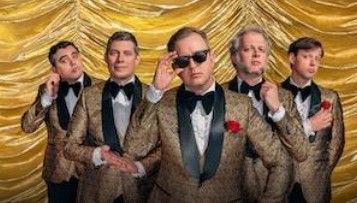 Vijf mannen in gouden smokingjasjes poseren op het podium; één van hen draagt een zonnebril.
