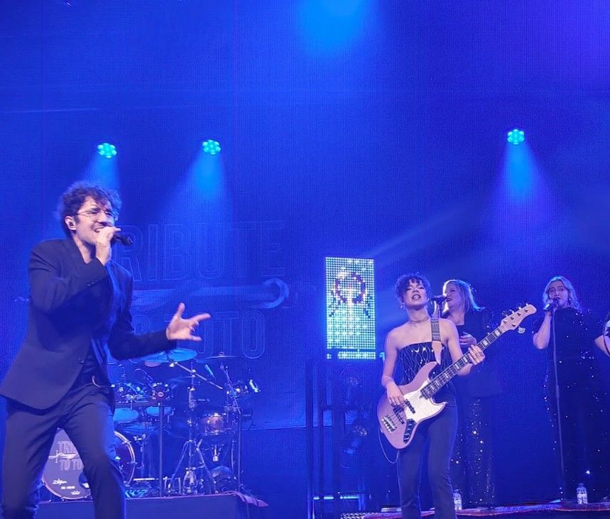 Band treedt op een podium op onder blauw licht; de leadzanger heeft zijn armen uitgestrekt.