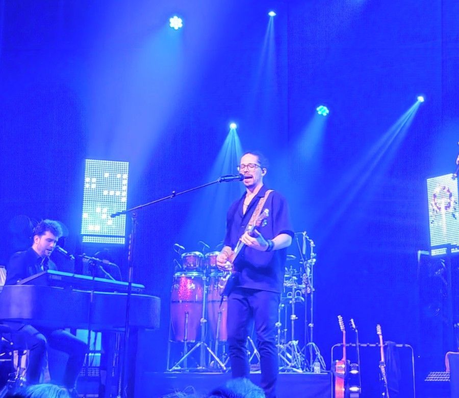 Band treedt op op een podium in blauw licht. De gitarist zingt terwijl hij speelt; de toetsenist staat links. Drums op de achtergrond.