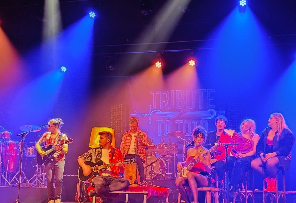 Band treedt op op een podium met blauwe en oranje podiumverlichting; op een spandoek staat