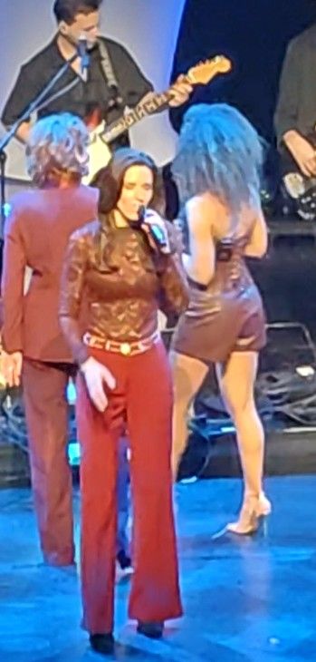 Een vrouw zingt op het podium in een microfoon, gekleed in een bordeauxrode top en rode broek.