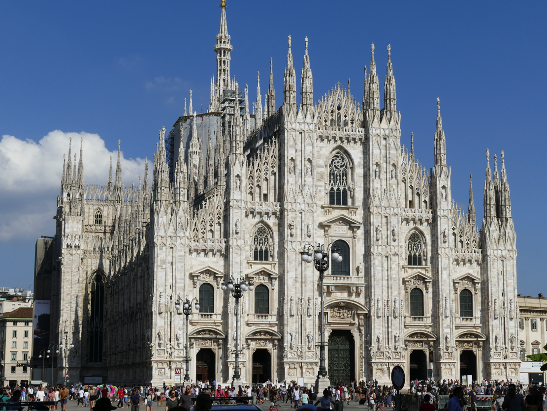 Duomo di Milano, een grote witte gotische kathedraal met ingewikkelde details, een blauwe lucht en mensen beneden.