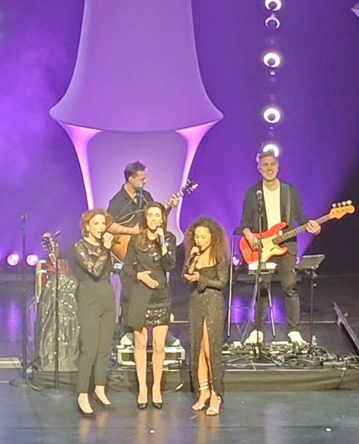 Drie vrouwen zingen op het podium met een band, gehuld in paars licht.