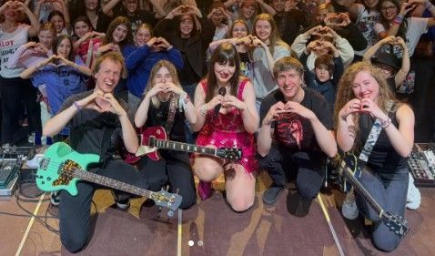 De bandleden poseren op het podium, lachend en hartjes makend, met gitaren en een juichend publiek achter hen.