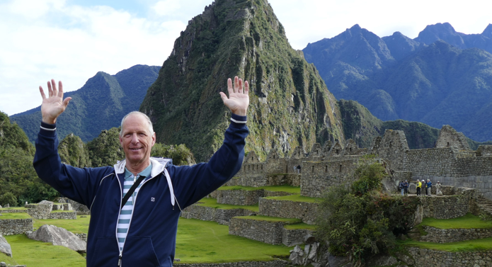 Man met opgeheven armen bij Machu Picchu. Oude stenen bouwwerken en een bergachtig decor vormen de achtergrond.