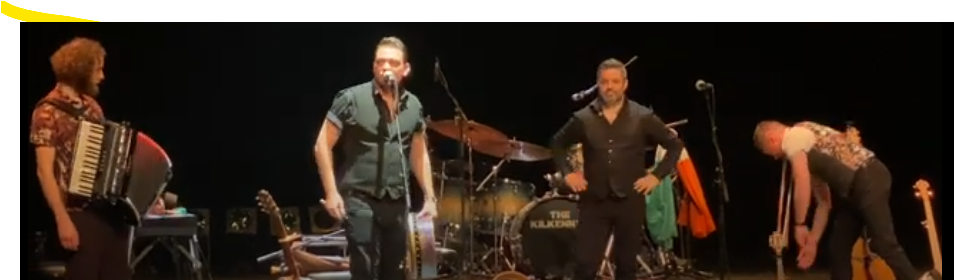 Een band die optreedt op een podium met een drummer, gitarist, zanger en accordeonist.