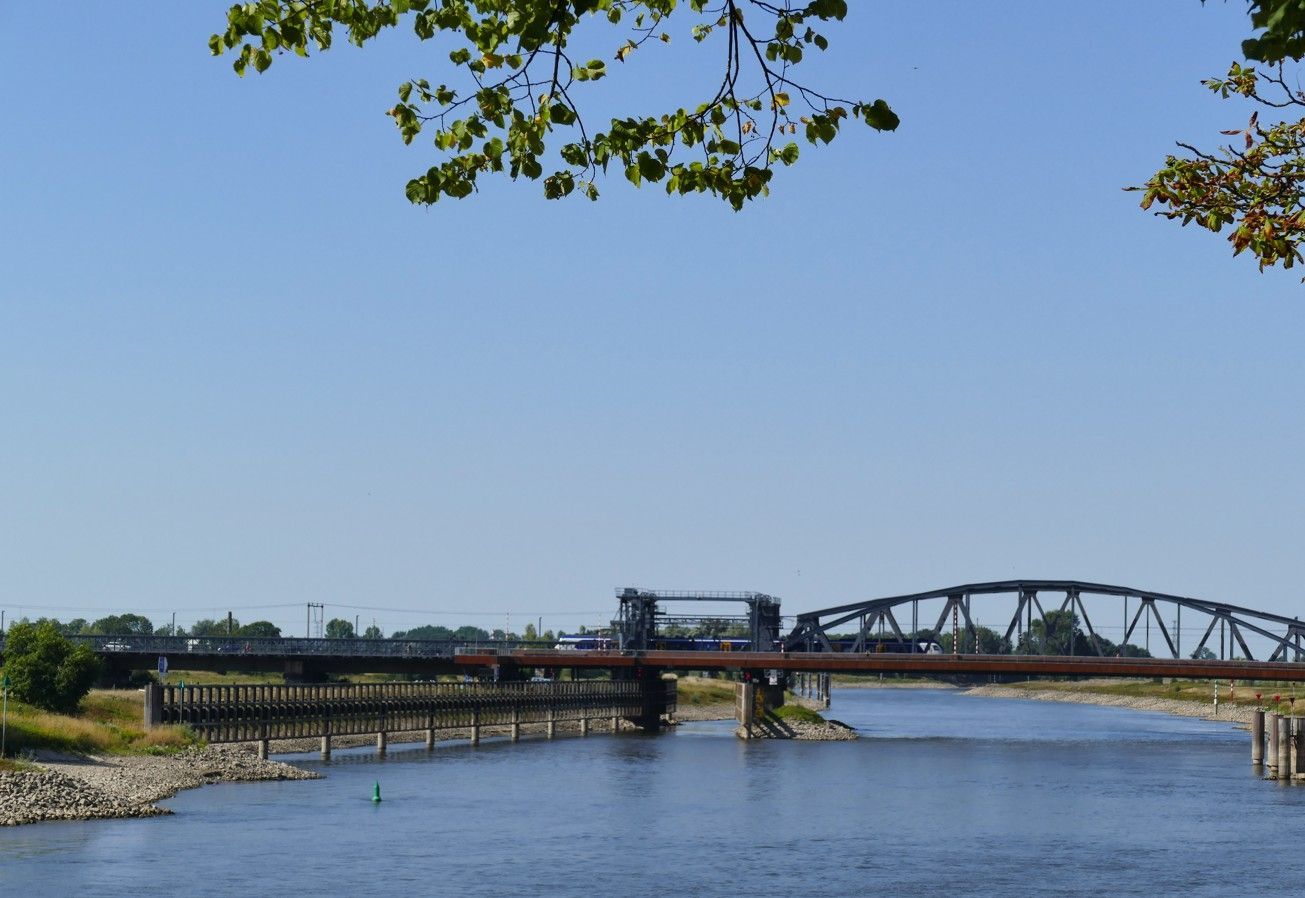Een rivier met een brug onder een helderblauwe hemel, omlijst door boomtakken.