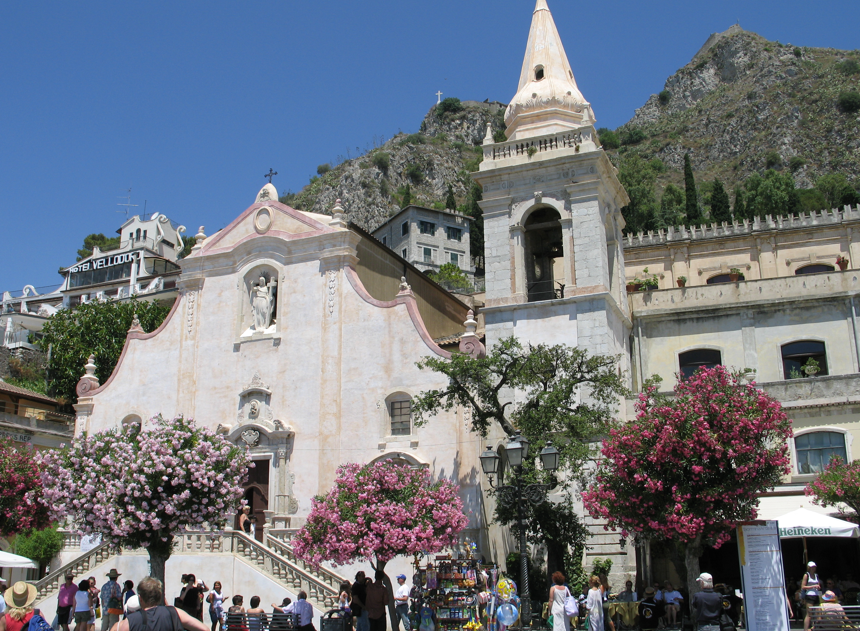 Kerk in Taormina, Italië, met klokkentoren en bloeiende roze bomen op een plein, waar mensen rondlopen.