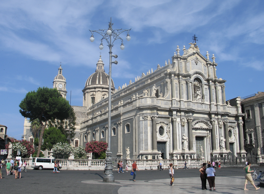 Exterieurbeeld van een grote, rijk versierde kerk in Catania, Italië, met mensen die op een zonnige dag over het plein lopen.