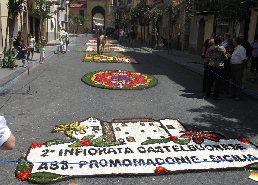 Kleurrijke bloemenkleden sieren een straat in Castelbuono, Sicilië, ter ere van een infiorata-festival; mensen kijken toe.