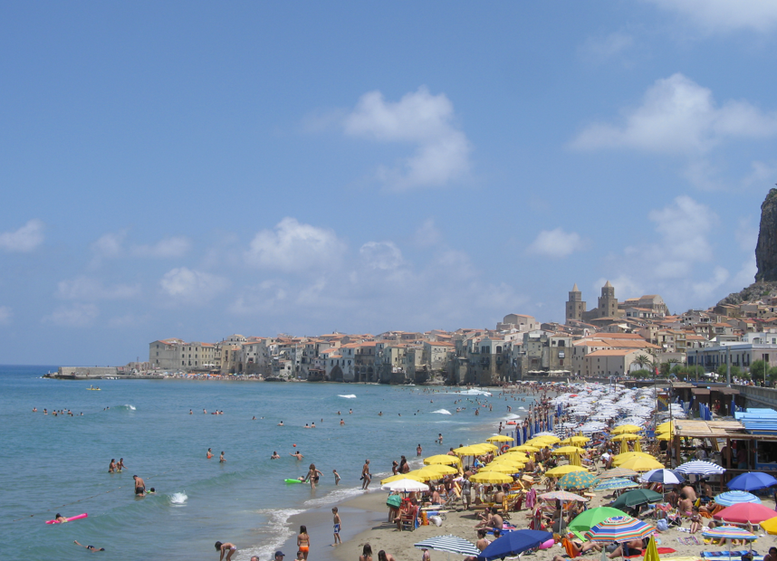 Strandscène in Cefalù, Italië, met kleurrijke parasols, gebouwen en mensen in het water en op het zand.